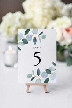 a table number card with eucalyptus leaves and white flowers in the background on a wooden stand