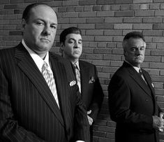 three men in suits and ties standing next to a brick wall with one man looking at the camera