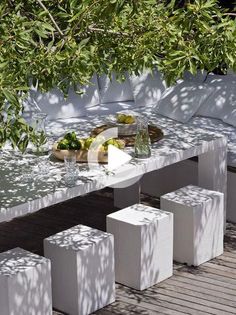 an outdoor table with fruit on it and white stools underneath the shade tree branches