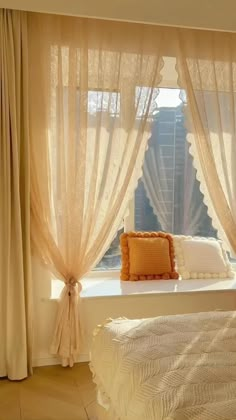 a white bed sitting next to a window covered in sheer curtains and an orange pillow