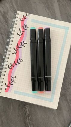 three black pens sitting on top of a notebook next to a pen and pencil sharpener