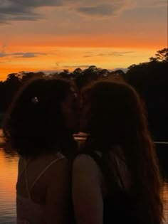 two women standing next to each other in front of a body of water at sunset