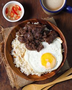 a bowl filled with rice, meat and eggs on top of a wooden table next to two cups of coffee