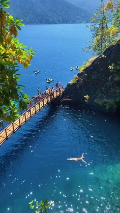 people are swimming in the blue water near a bridge that is surrounded by trees and mountains