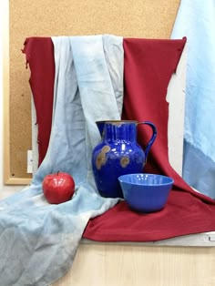 a blue pitcher, bowl and an apple on a red cloth next to a cork board