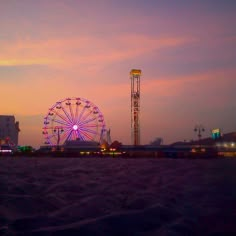 a ferris wheel sitting on top of a sandy beach next to tall buildings at dusk