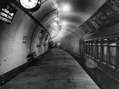 a black and white photo of a subway station with a clock hanging from the ceiling