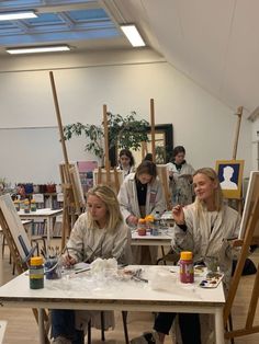 three women sitting at a table painting in an art room with easels and paintings