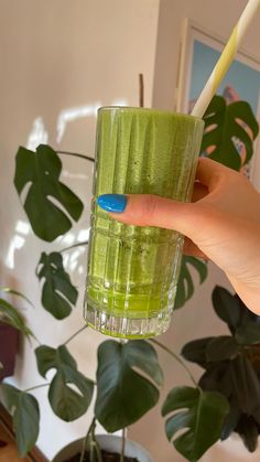 a hand holding a green drink with a yellow straw in it and a plant behind it