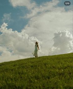 a woman in a white dress is standing on a grassy hill and looking up at the sky