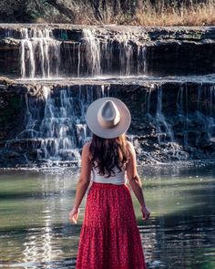 a woman in a red dress and hat standing by a waterfall looking at the water