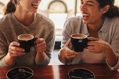 two women sitting at a table with coffee cups in their hands, laughing and looking into the camera