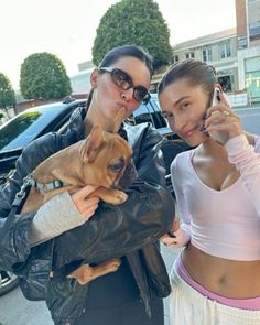 two women standing next to each other while one holds a dog on her cell phone