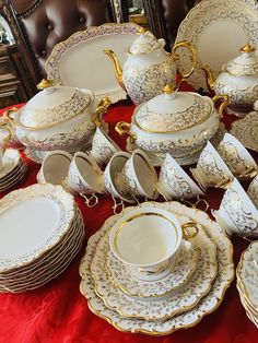 a table topped with lots of white and gold china dishes on top of a red table cloth