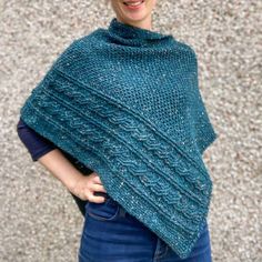 a woman wearing a green knitted ponchy standing in front of a stone wall