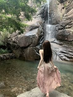 a woman standing in front of a waterfall with her back to the camera, looking down