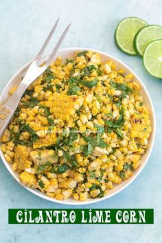 a bowl filled with corn, cilantro and limes next to two forks