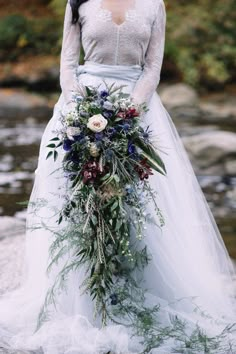 a woman in a wedding dress holding a bouquet with flowers on the side, standing next to a river