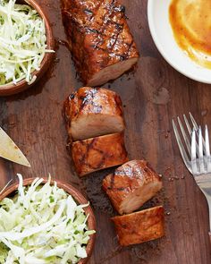 some meat and coleslaw are on a cutting board next to two bowls with silverware