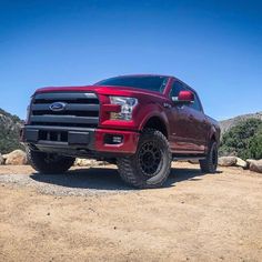 a red truck parked on top of a dirt field next to rocks and trees in the background