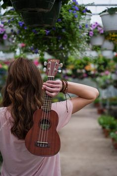 a woman holding a ukulele in her hand and looking at flowers hanging from the ceiling