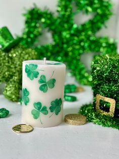 a white candle with green shamrocks on it next to some gold coins and trees