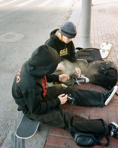 a person sitting on the ground with a skateboard in front of him and another person standing next to them