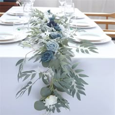 the table is set with white and blue flowers, greenery, and silverware