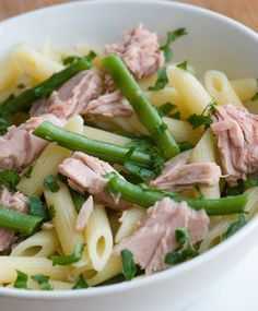 a white bowl filled with pasta and meat on top of a wooden table next to green beans