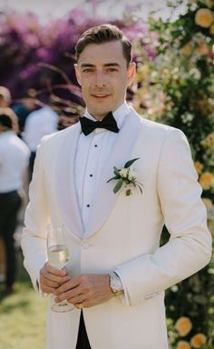 a man in a white tuxedo holding a wine glass and looking at the camera