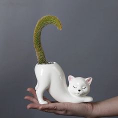 a hand holding a white ceramic cat with a green plant in it's mouth