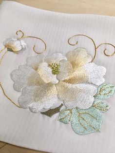 a white flower with green leaves is on a table cloth that has been embroidered onto it