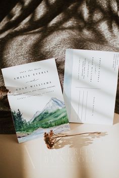 two wedding cards sitting on top of a table next to each other with trees and mountains in the background