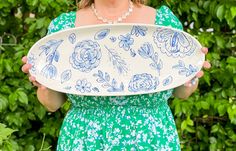 a woman holding a plate with blue flowers on it in front of green bushes and shrubbery