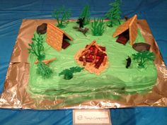 a cake that is shaped like a camp site with trees and grass on it, sitting on a blue table cloth