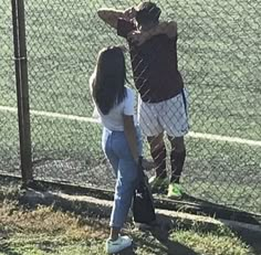 two people standing next to each other near a fence and grass area with a tennis court in the background