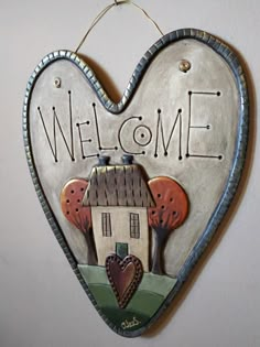 a heart shaped welcome sign hanging from the side of a wall with a house and trees painted on it