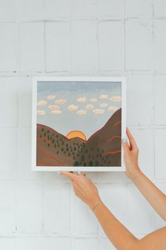 a woman holding up a painting with mountains in the background and clouds in the sky