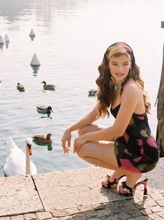 a woman kneeling down next to a body of water with ducks in the lake behind her