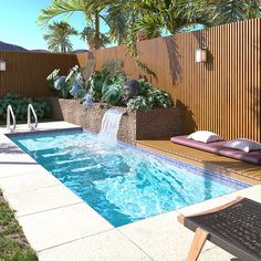 a small pool with a waterfall in the middle and lounge chairs around it, next to a wooden fence