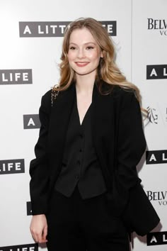 a woman standing in front of a white wall wearing a black suit and smiling at the camera