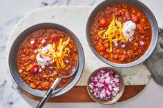 two bowls filled with chili and cheese on top of a cutting board next to a bowl of onions