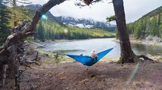 a person sitting in a blue hammock near a lake and trees with mountains in the background