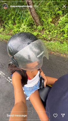 a woman riding on the back of a motorcycle down a road next to lush green trees