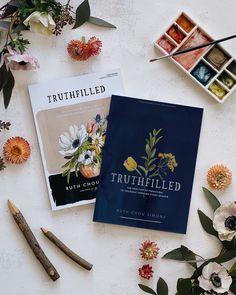 two books on the table surrounded by flowers and other items that include paintbrushes