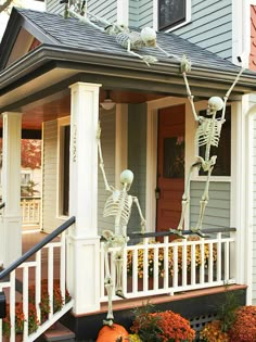 two skeleton statues on the porch of a house with pumpkins and flowers in front