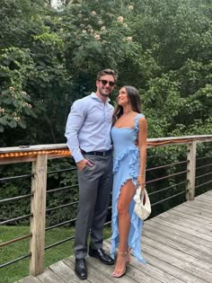 a man and woman standing on a wooden deck in front of some trees, posing for the camera