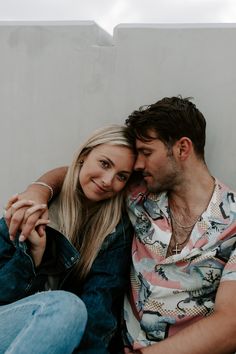 a man and woman sitting next to each other in front of a wall with their arms around each other