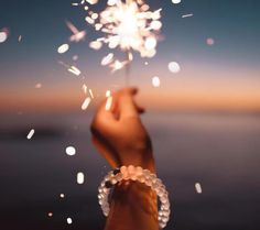 someone holding a sparkler in their hand with the sun setting on the water behind them