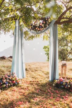 an outdoor ceremony set up with blue drapes and flowers on the ground under a tree